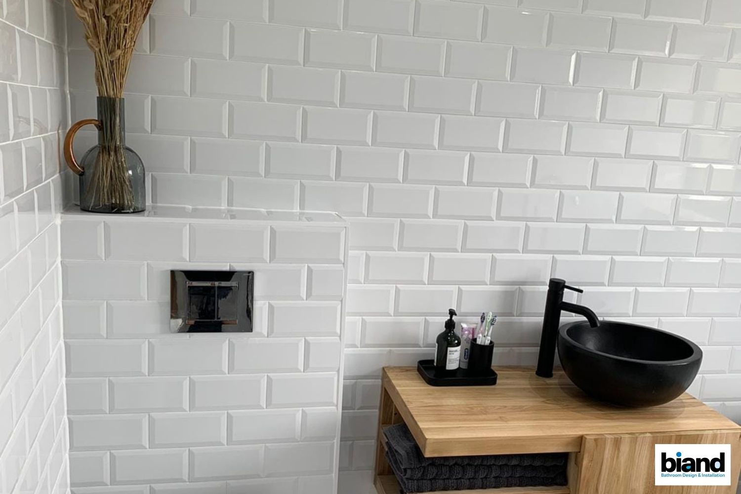 White subway tile bathroom with black sink, wooden vanity, and decorative vase.