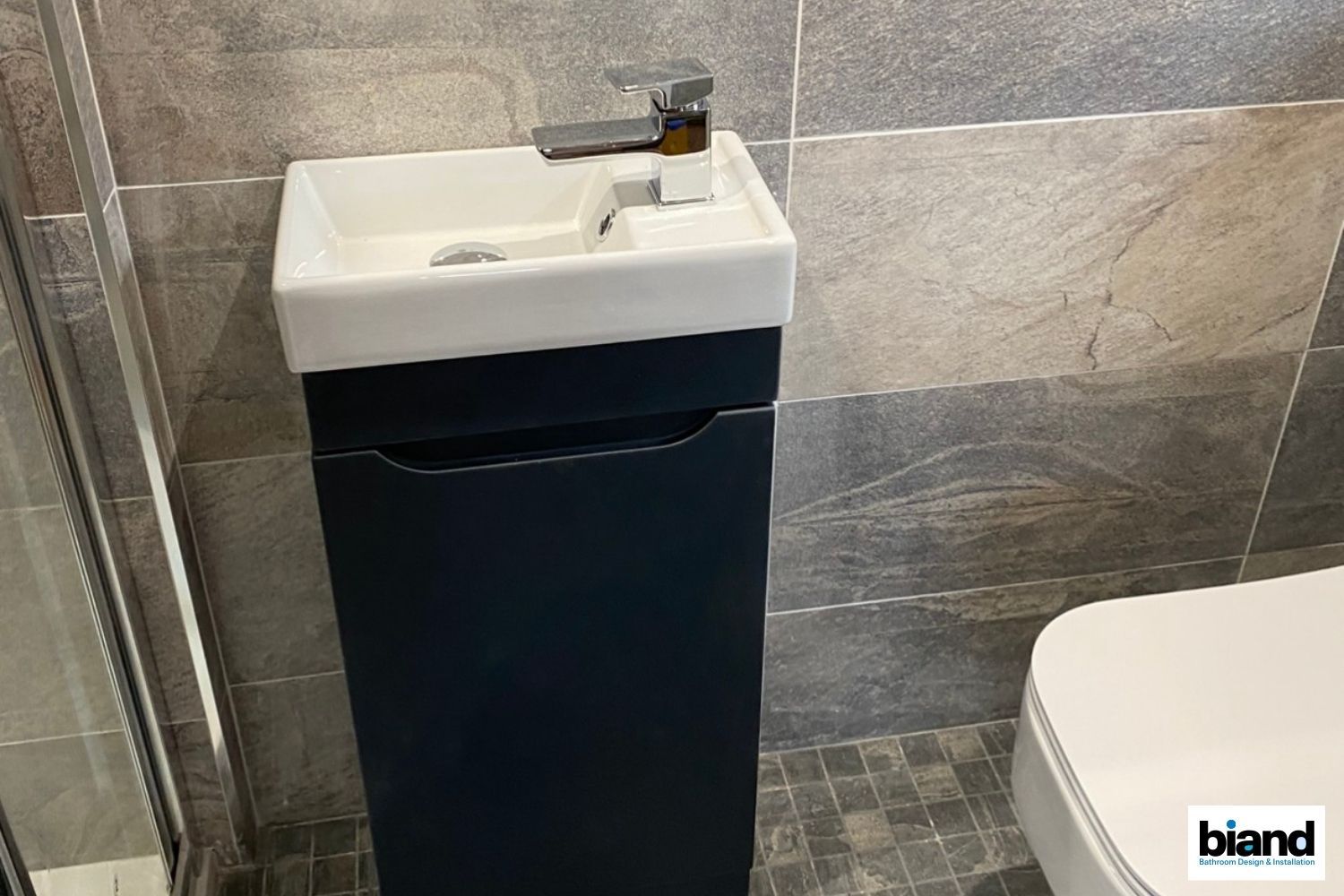 Narrow navy vanity with white sink in a bathroom; gray tile walls and small floor tiles.
