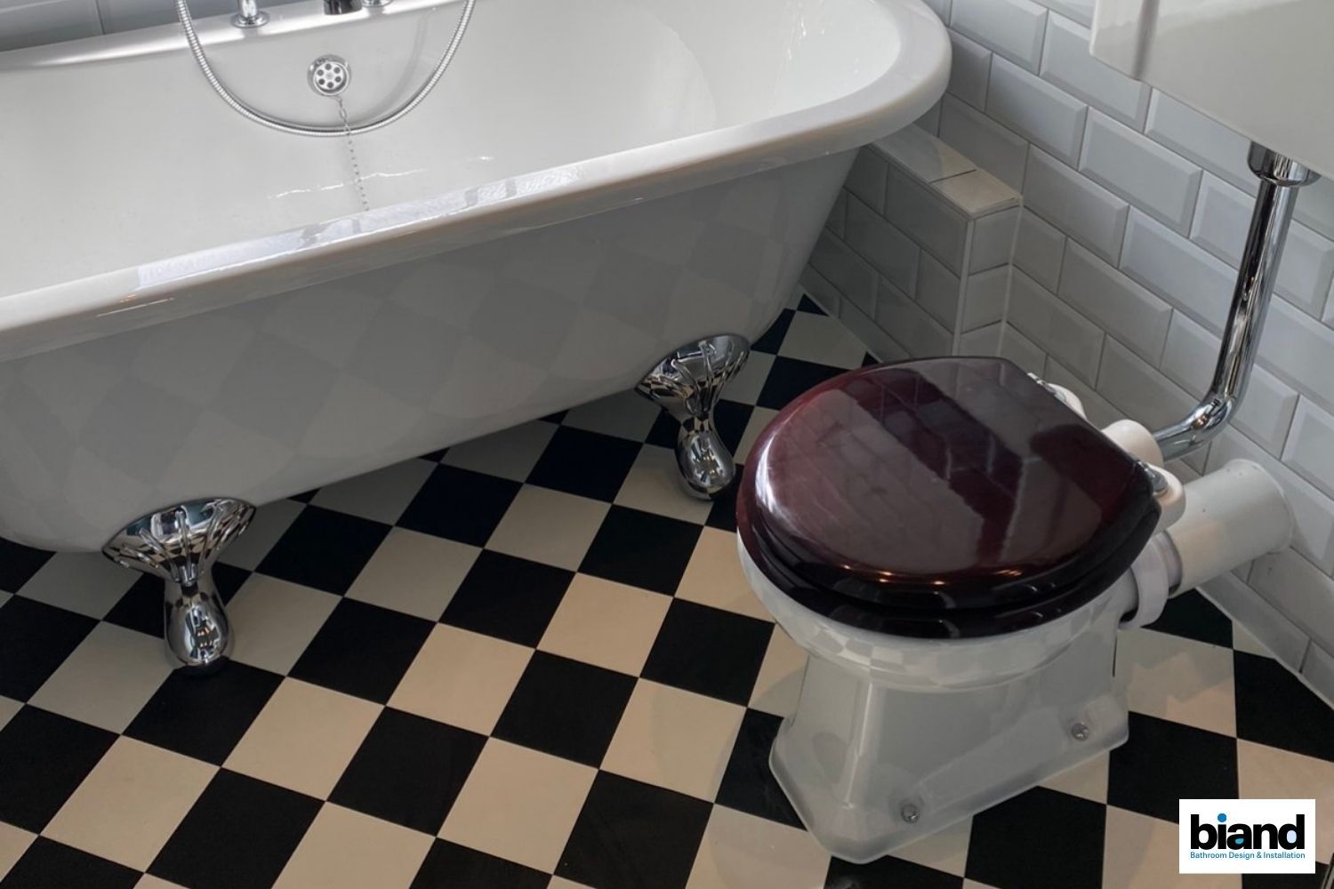 Claw-foot tub, toilet with wooden seat in a bathroom. Black and white checkered floor.