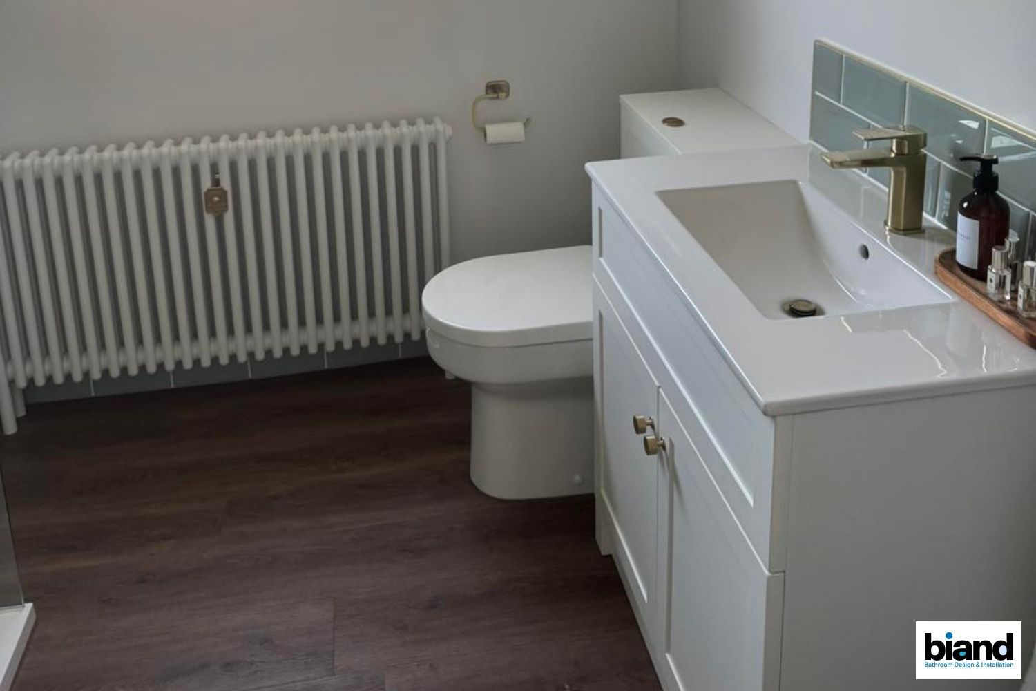 White bathroom with a sink, toilet, radiator, and wood-look floor.