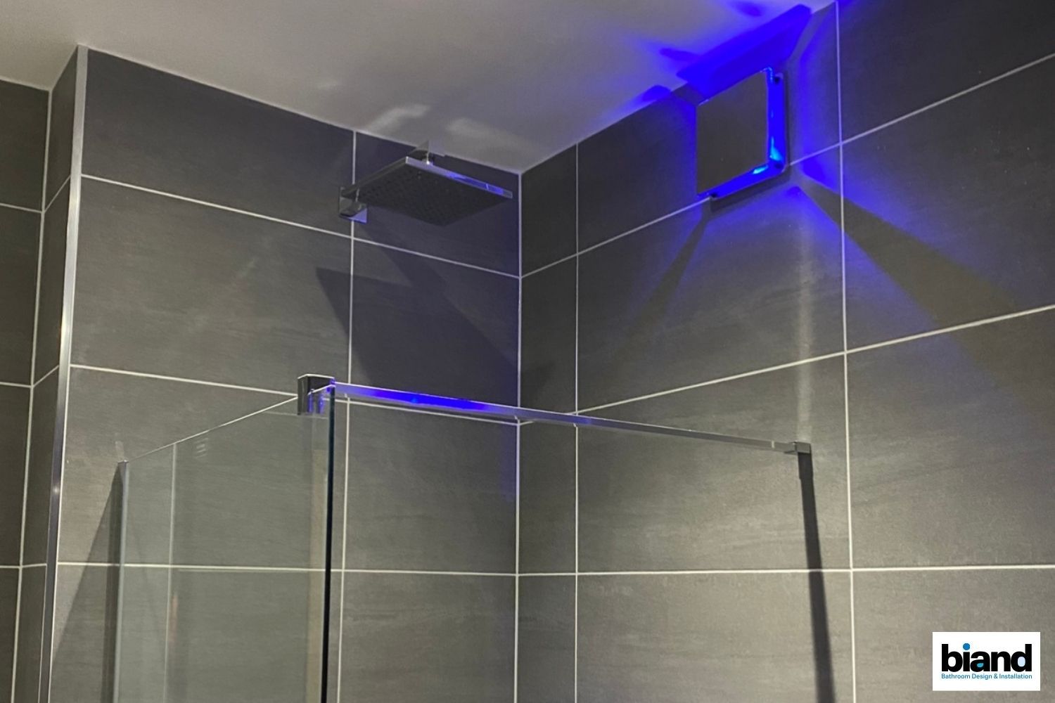 Bathroom corner with dark gray tiles, a shower head, glass door, and blue LED lighting.