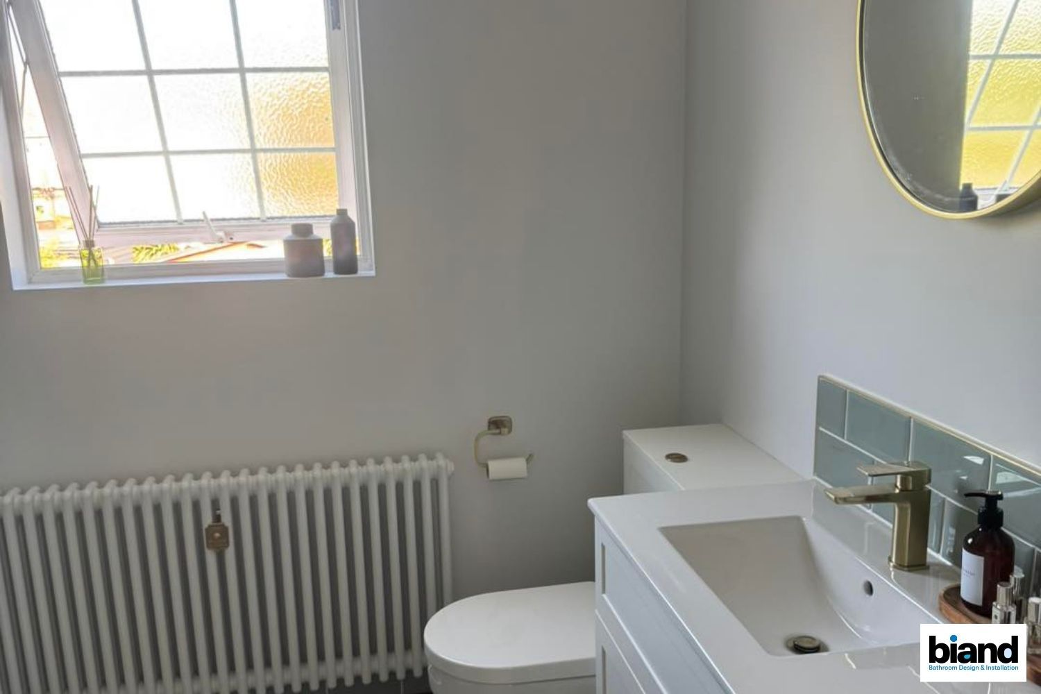 Small, light-filled bathroom with toilet, sink, radiator, and a window with stained glass.
