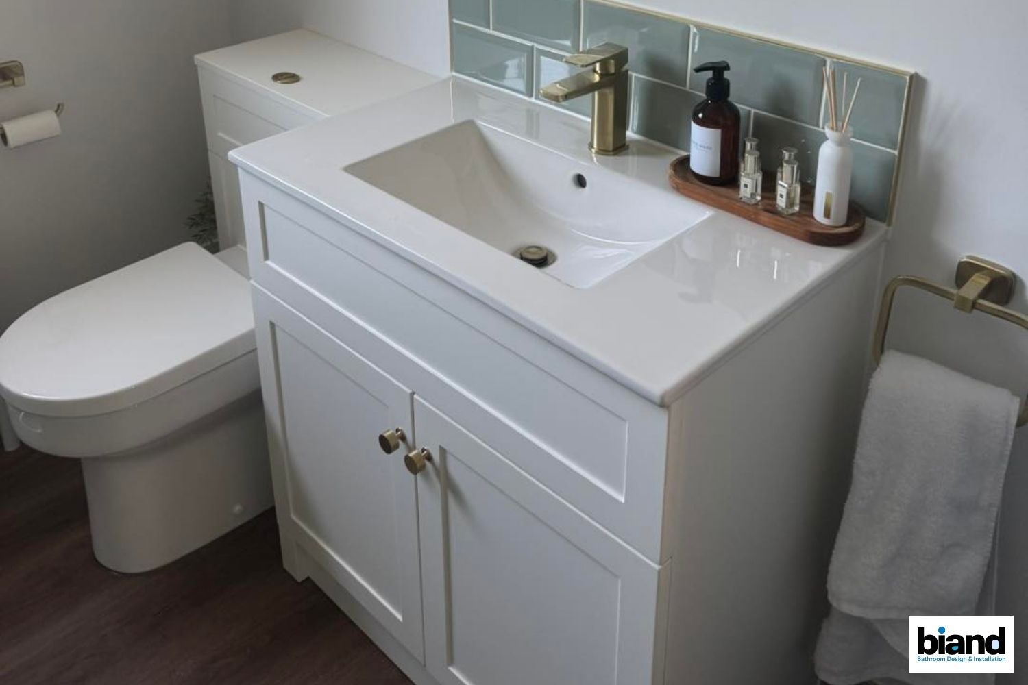 White bathroom vanity and toilet, gold fixtures, teal backsplash, wooden shelf with toiletries.