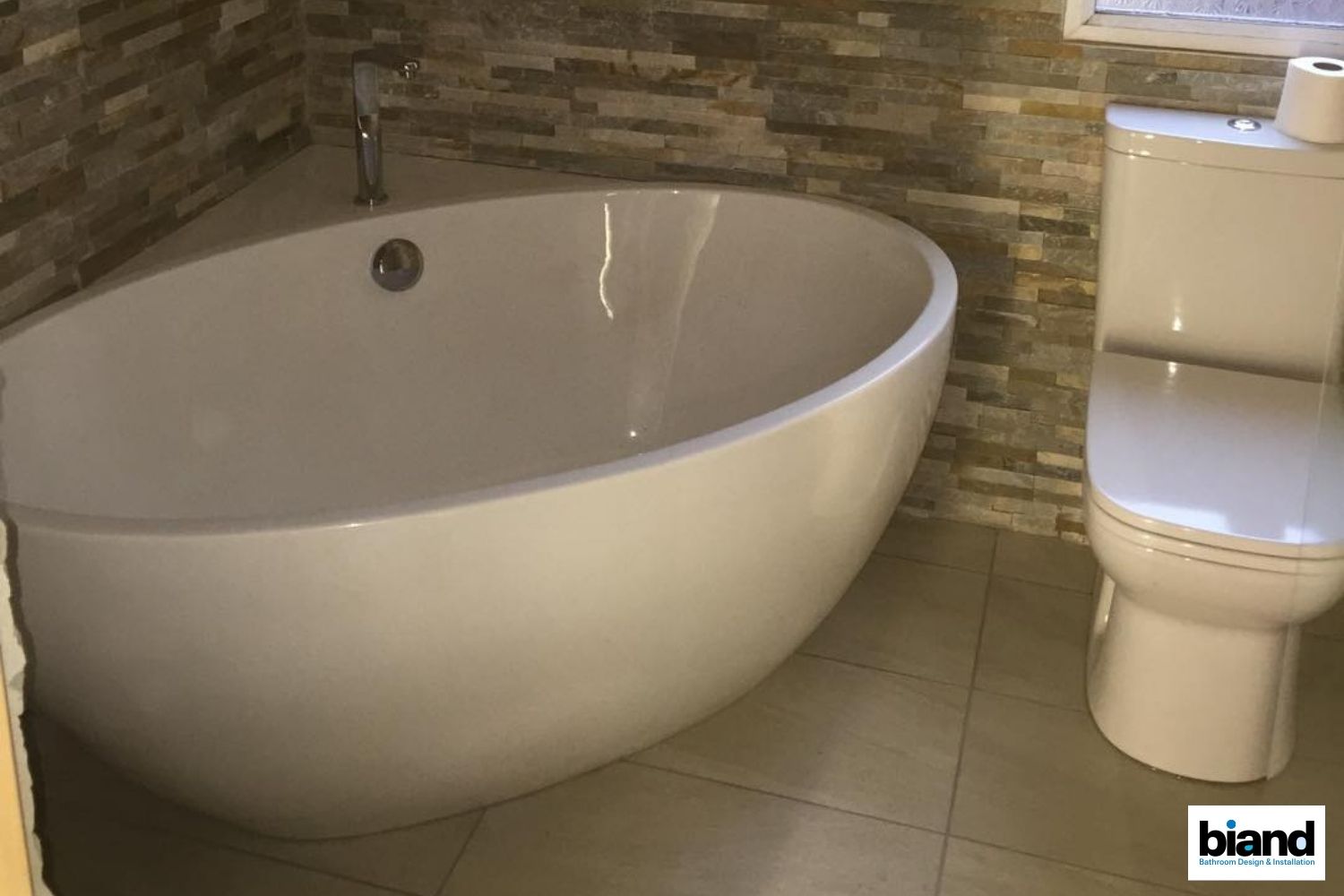 Oval white bathtub next to a white toilet in a bathroom with stone tile accents.