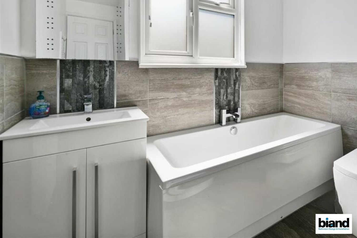 White bathroom with tub, sink, and cabinet. Beige tiled wall, white window, and toilet.