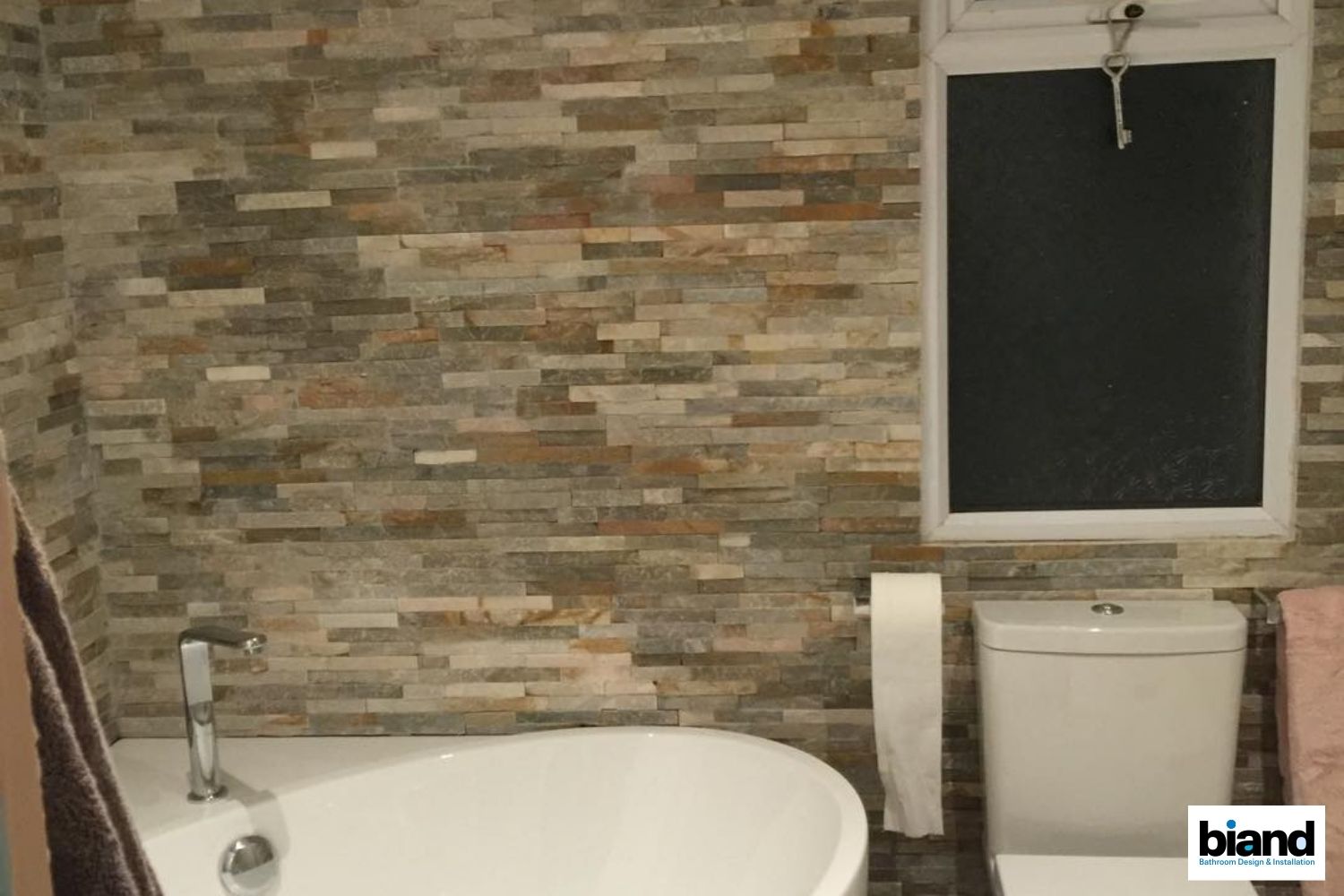 Bathroom with stone tile wall, bathtub, toilet, and window.