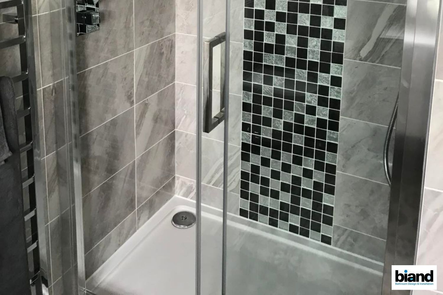 Shower stall with glass door, gray tiles, black and white mosaic accent, and chrome fixtures.
