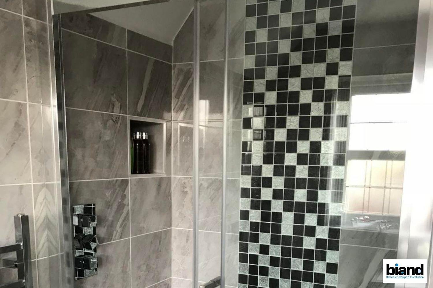 A modern bathroom shower with gray marble tiles, black and white mosaic, and glass doors.