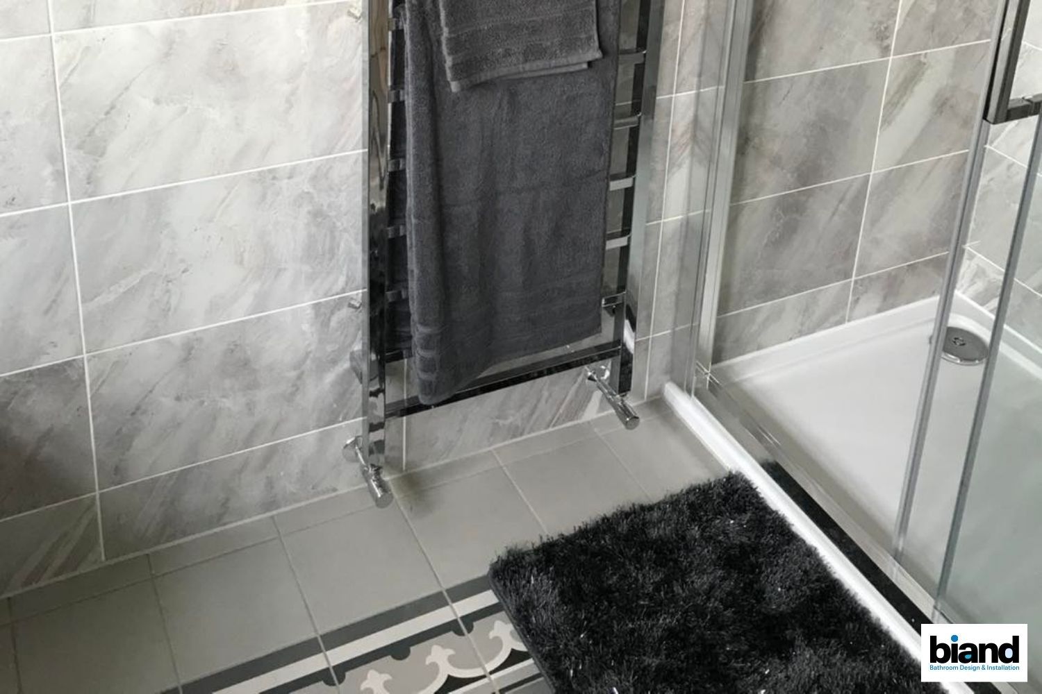 Bathroom with gray tiled walls, chrome towel rack with gray towels, and a black rug.