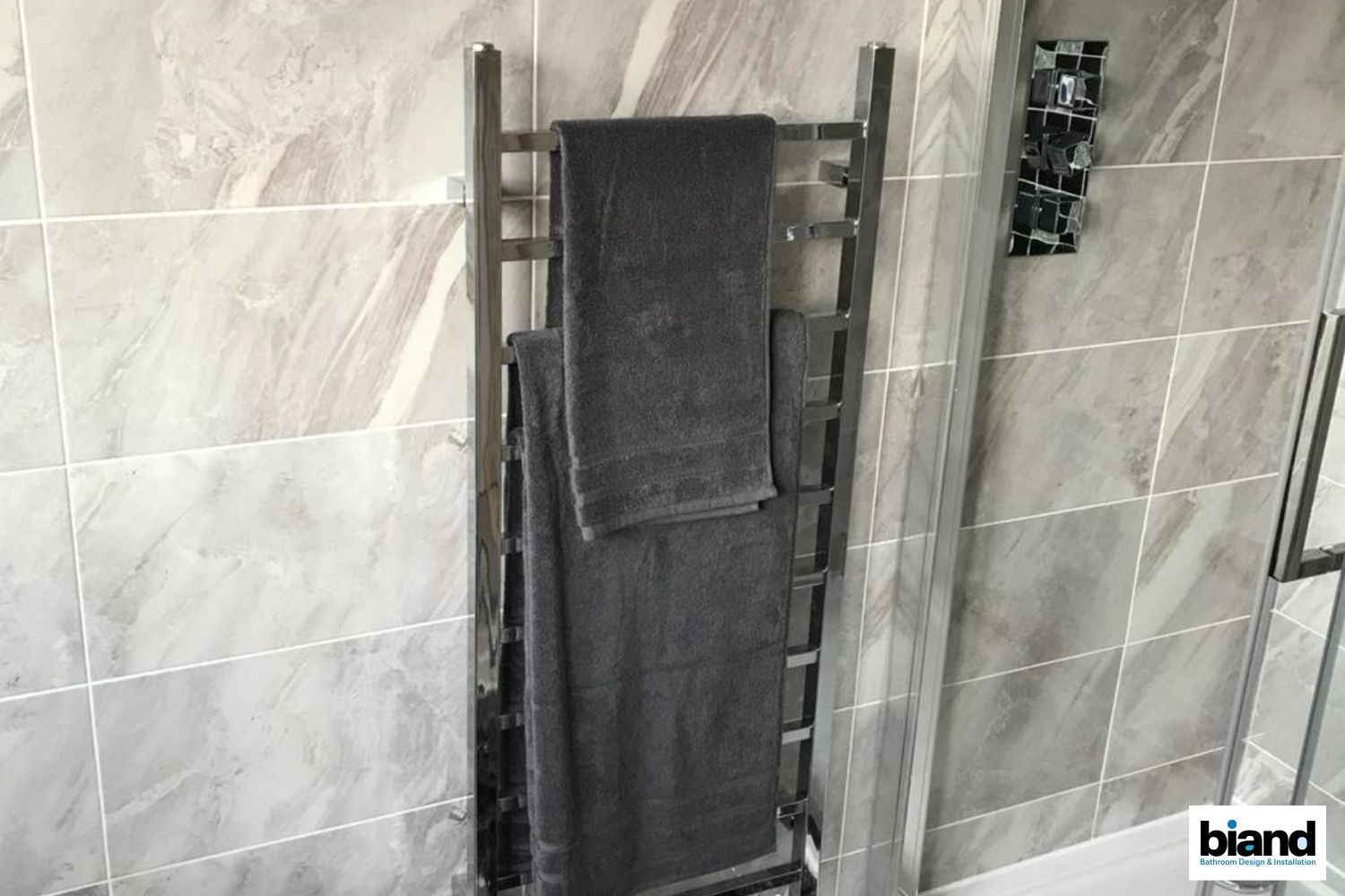 Towel warmer with two gray towels hanging on it, set against a tiled bathroom wall.