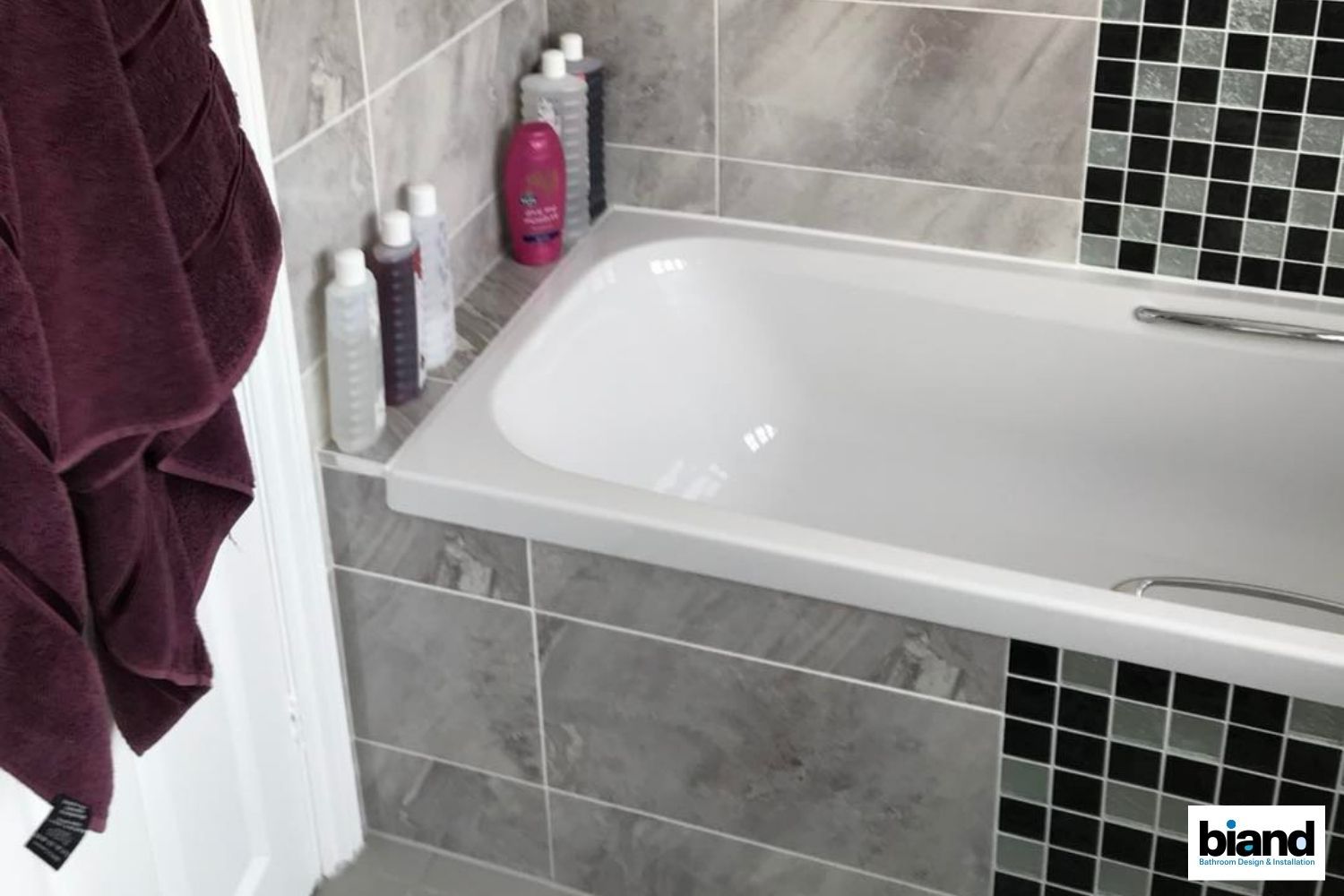 Bathtub in a tiled bathroom; towels, shampoo bottles on the ledge, grey, black and white mosaic accents.