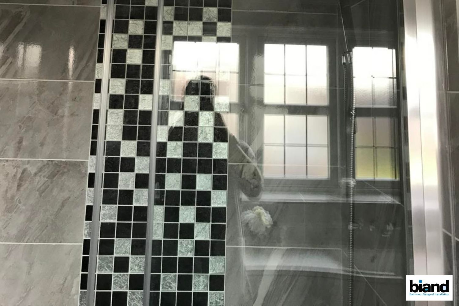 Shower interior with checkered tile strip and glass partition reflecting a window and a person.