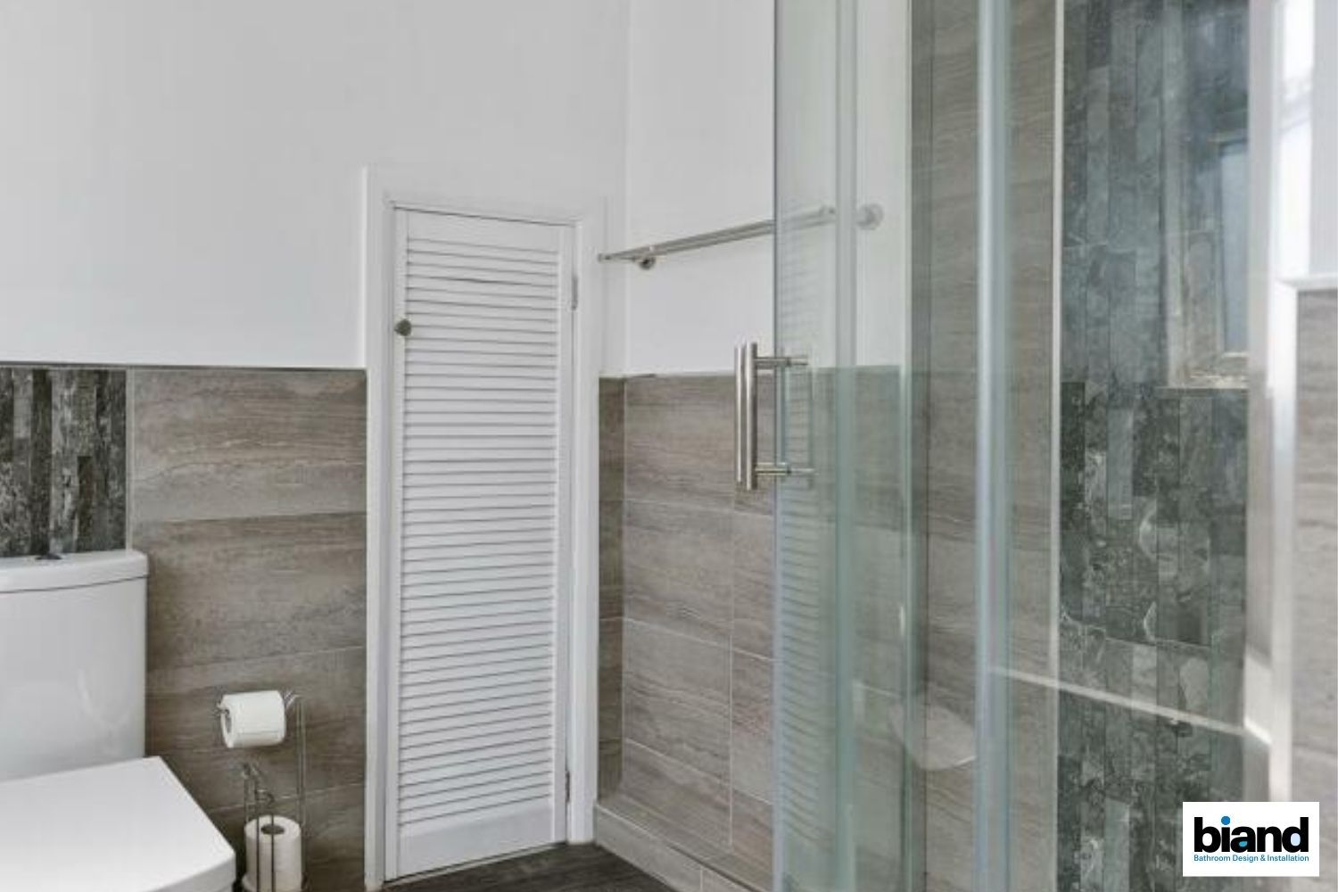 Bathroom with a white toilet, shower, and a white slatted door, gray tile walls.