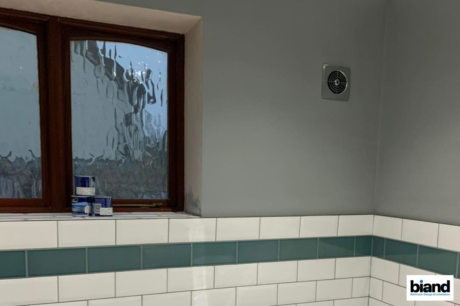Bathroom with white and teal tiles, window with textured glass, and a ventilation fan.