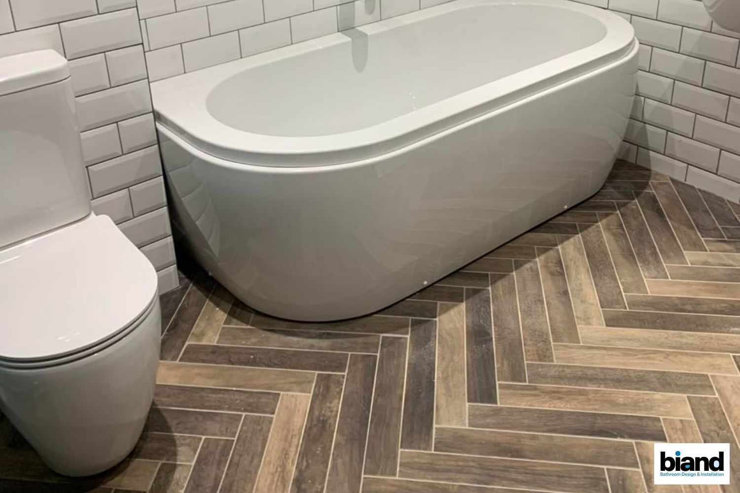 Bathroom with a white tub and toilet on herringbone wood-look tile flooring, white subway tile walls.
