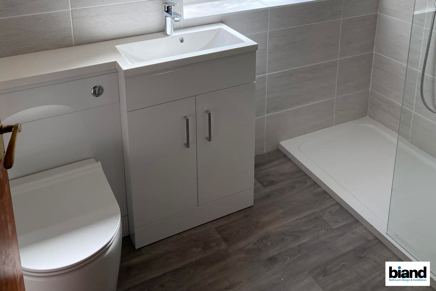 Small bathroom with white toilet, sink cabinet, and shower. Wood-look flooring and gray tiled walls.