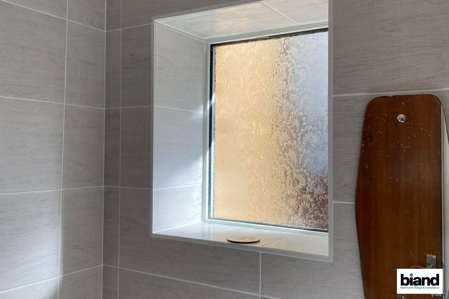 Bathroom with gray tiled walls, window with frosted glass, and wooden shelf.