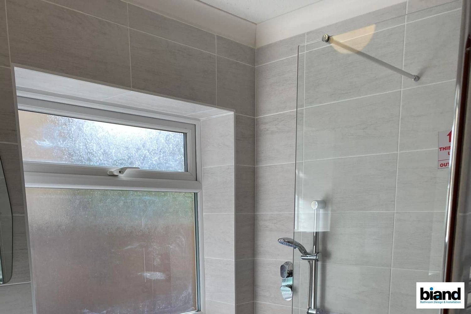 Gray-tiled bathroom with shower and window. Glass shower door, chrome fixtures. White ceiling, frosted window glass.