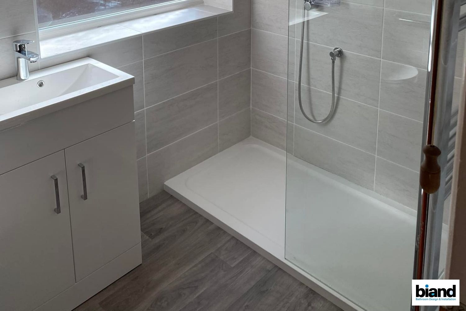 Bathroom with white shower, sink, and cabinet. Gray tiled walls, wood-look flooring, and window.