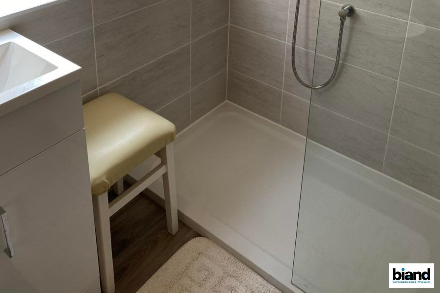 Small bathroom with a shower, a sink, and a stool. Beige tiles and a white shower tray.