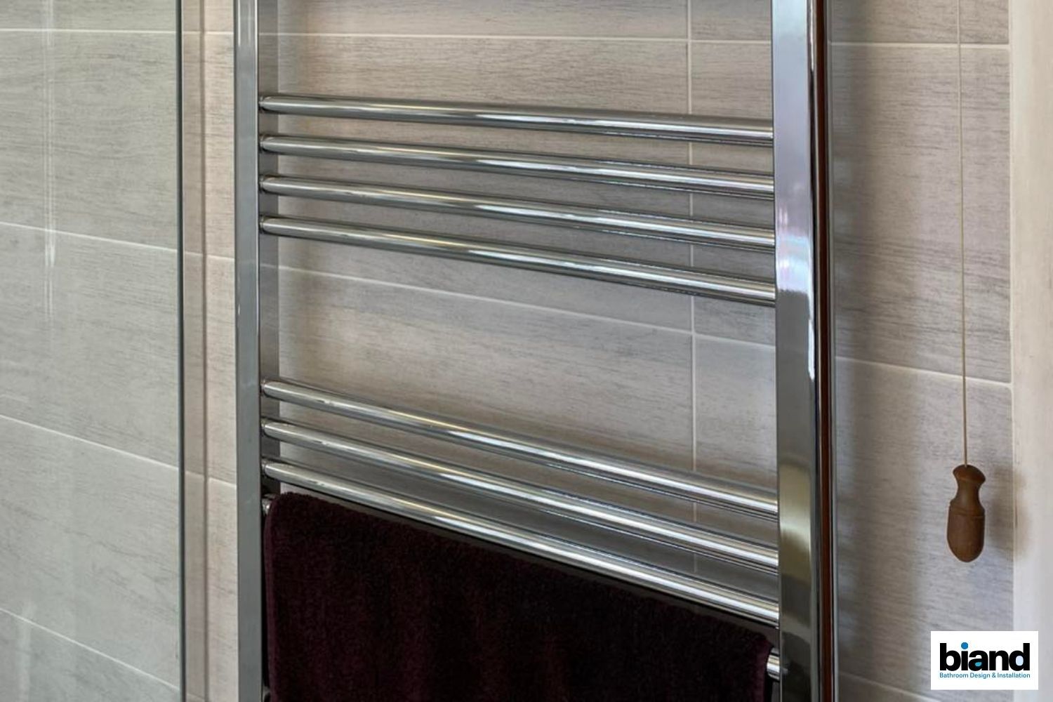 Chrome towel warmer in a bathroom; dark towel hanging, light gray tile wall, pull cord.