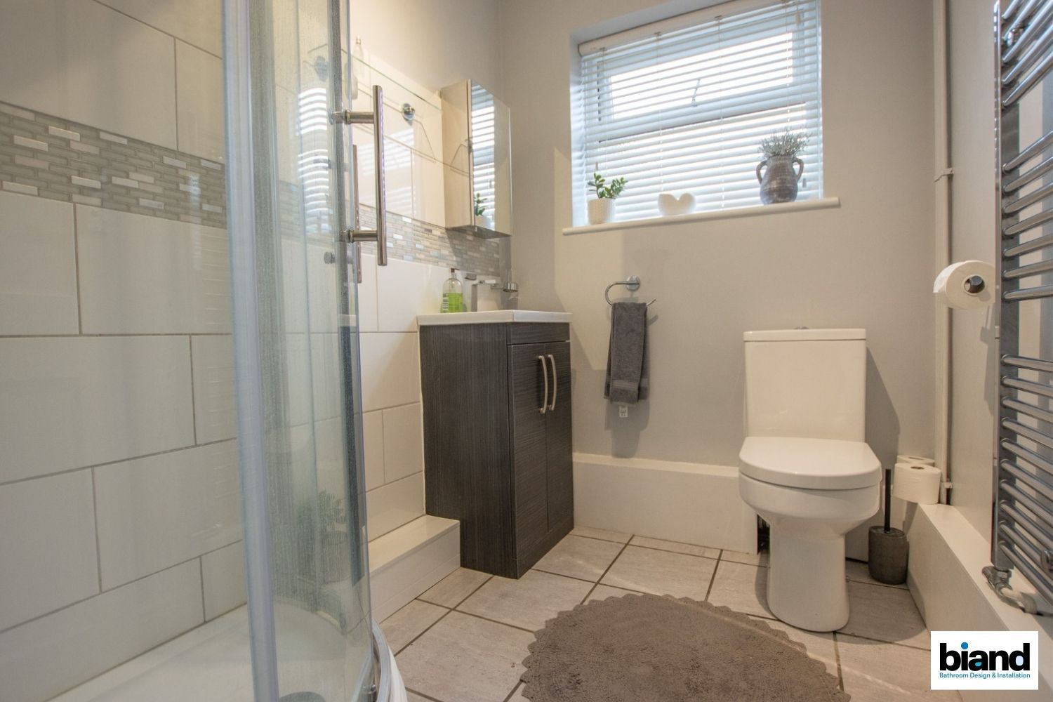 Bathroom with shower, sink, toilet, and window. Gray and white tiles, gray vanity.