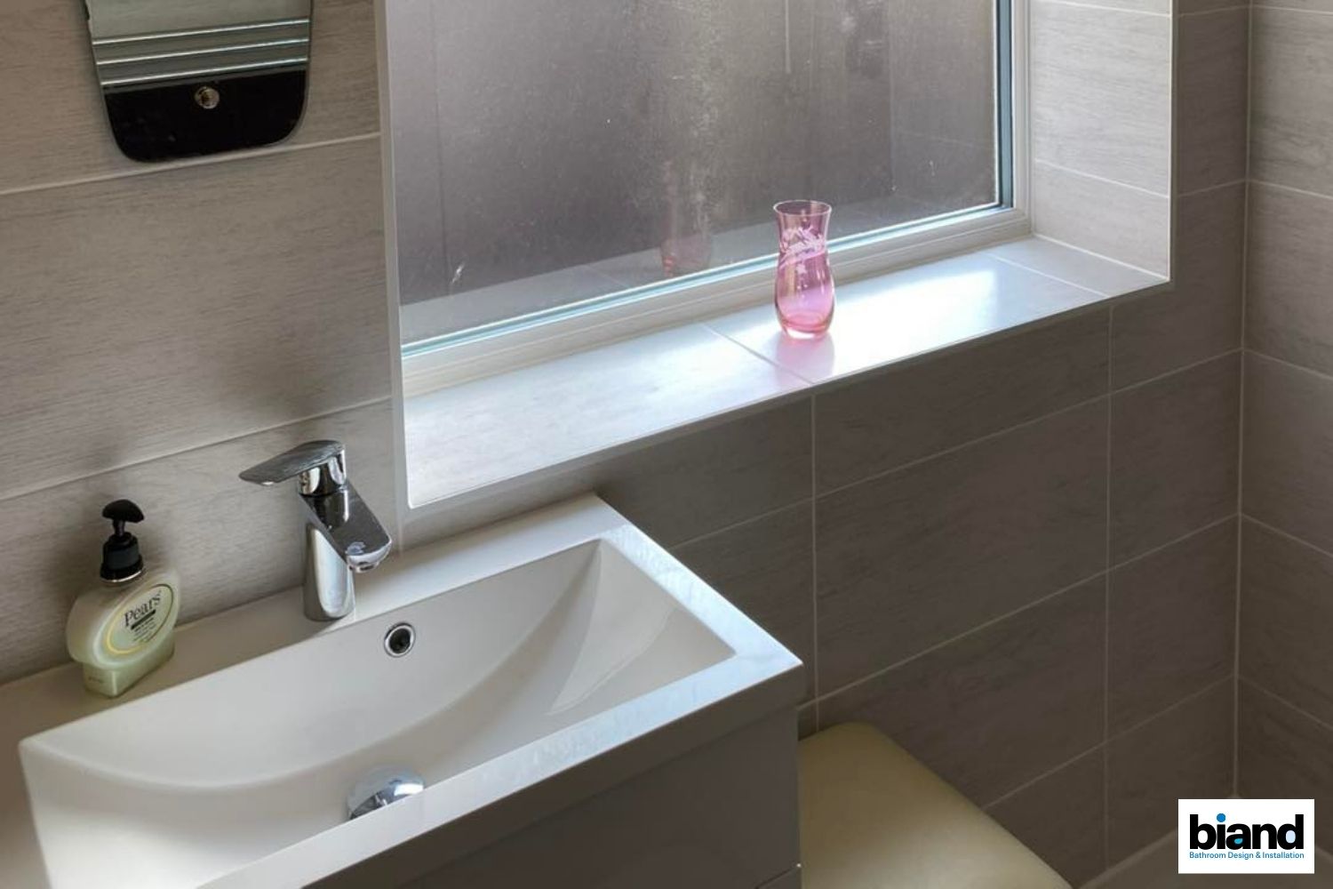 Bathroom sink and counter with a window and a pink vase on the windowsill. Light brown tiles and beige tones.