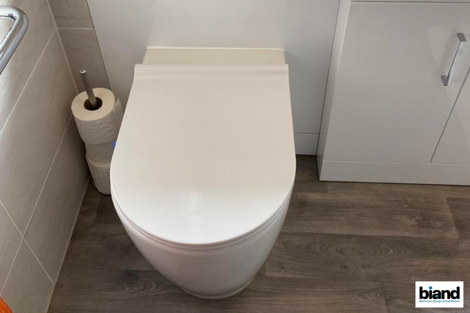 White toilet in a small bathroom next to a toilet paper holder and cabinet on wood-look flooring.