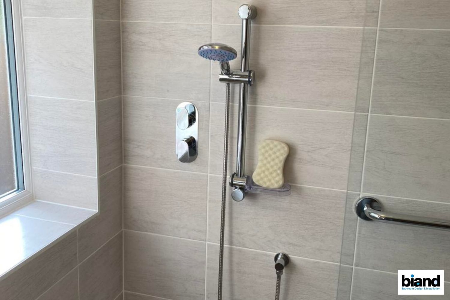 Shower with chrome fixtures, beige tile walls, sponge, and a grab bar.