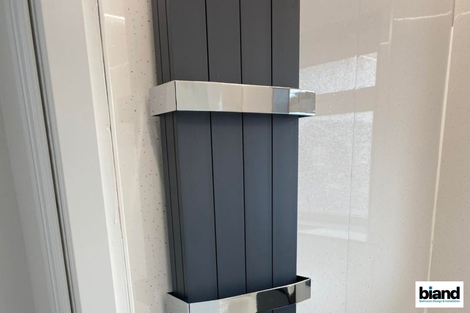 Gray vertical radiator with chrome accents mounted on a white wall.