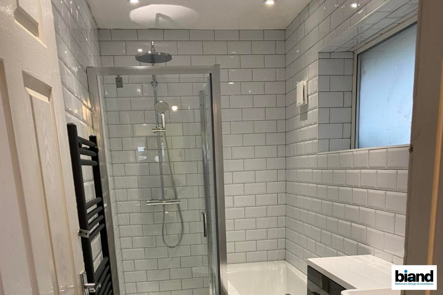 Small, white-tiled bathroom with a shower, window, and black towel rack.