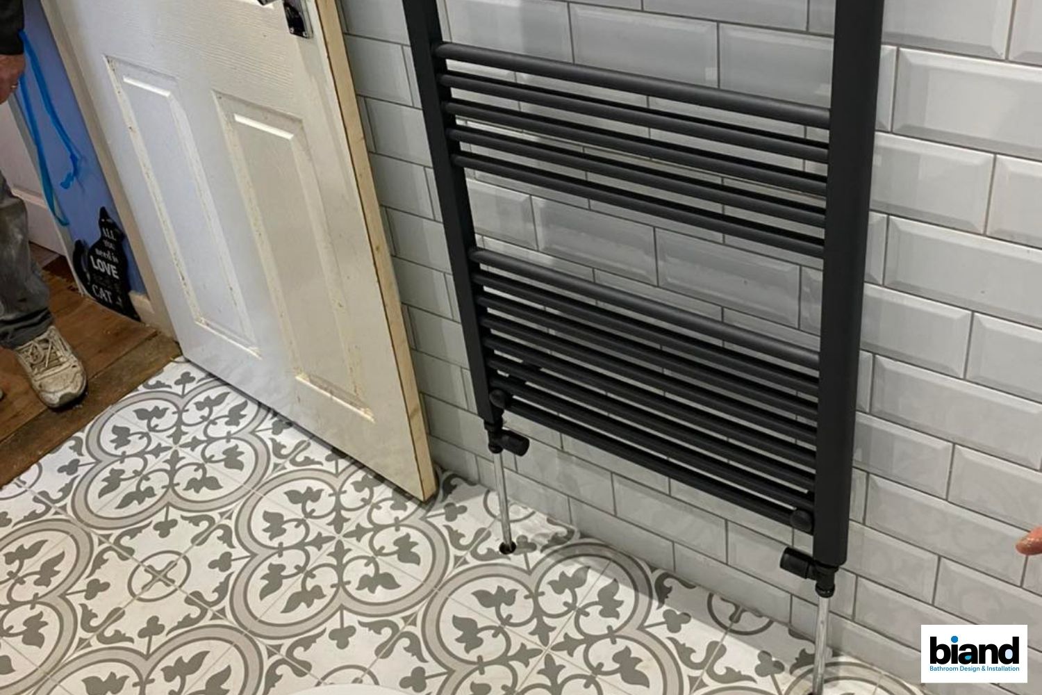 Black towel radiator leaning against a white-tiled wall in a bathroom. Ornate patterned floor and open door.