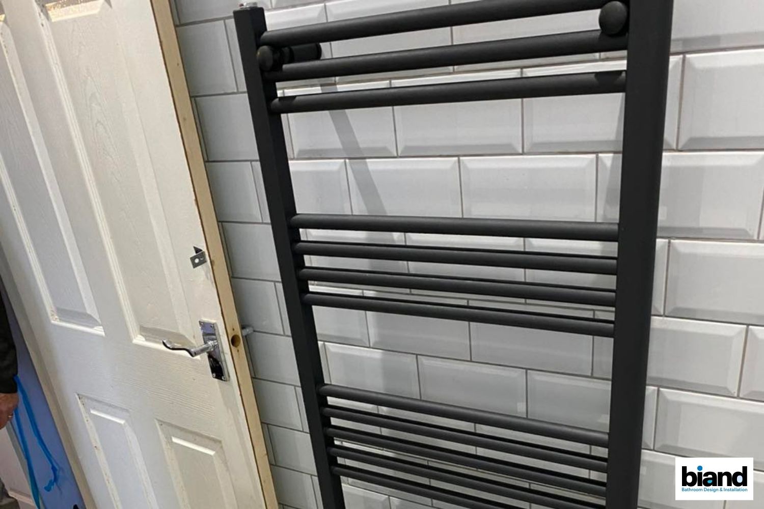 Black towel warmer on a white tiled wall next to a white door.