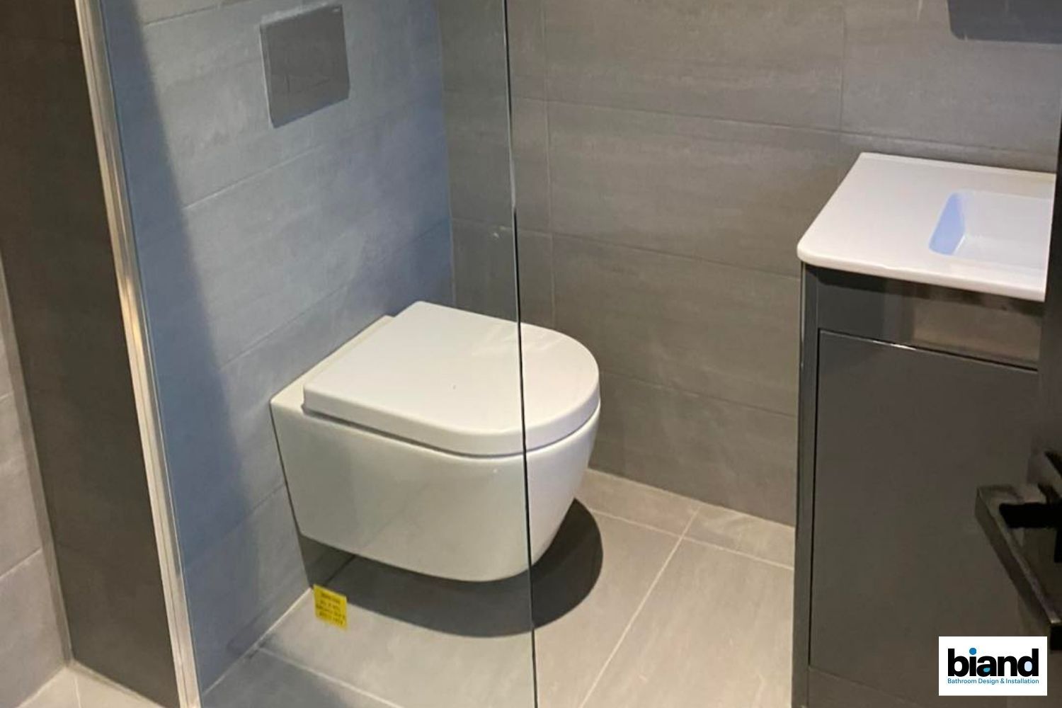 Modern bathroom with a wall-mounted toilet, grey tiles, and a vanity with a white top.