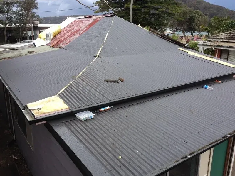 A Man in a Red Shirt is Standing on the Roof of a Building — Coast Metal Roof & Gutter In Morisset, NSW