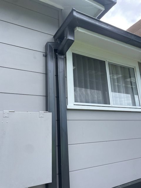 A black guttering downpipe stretching from the ground to a roof on a grey house — Coast Metal Roof & Gutter In Tumbi Umbi, NSW