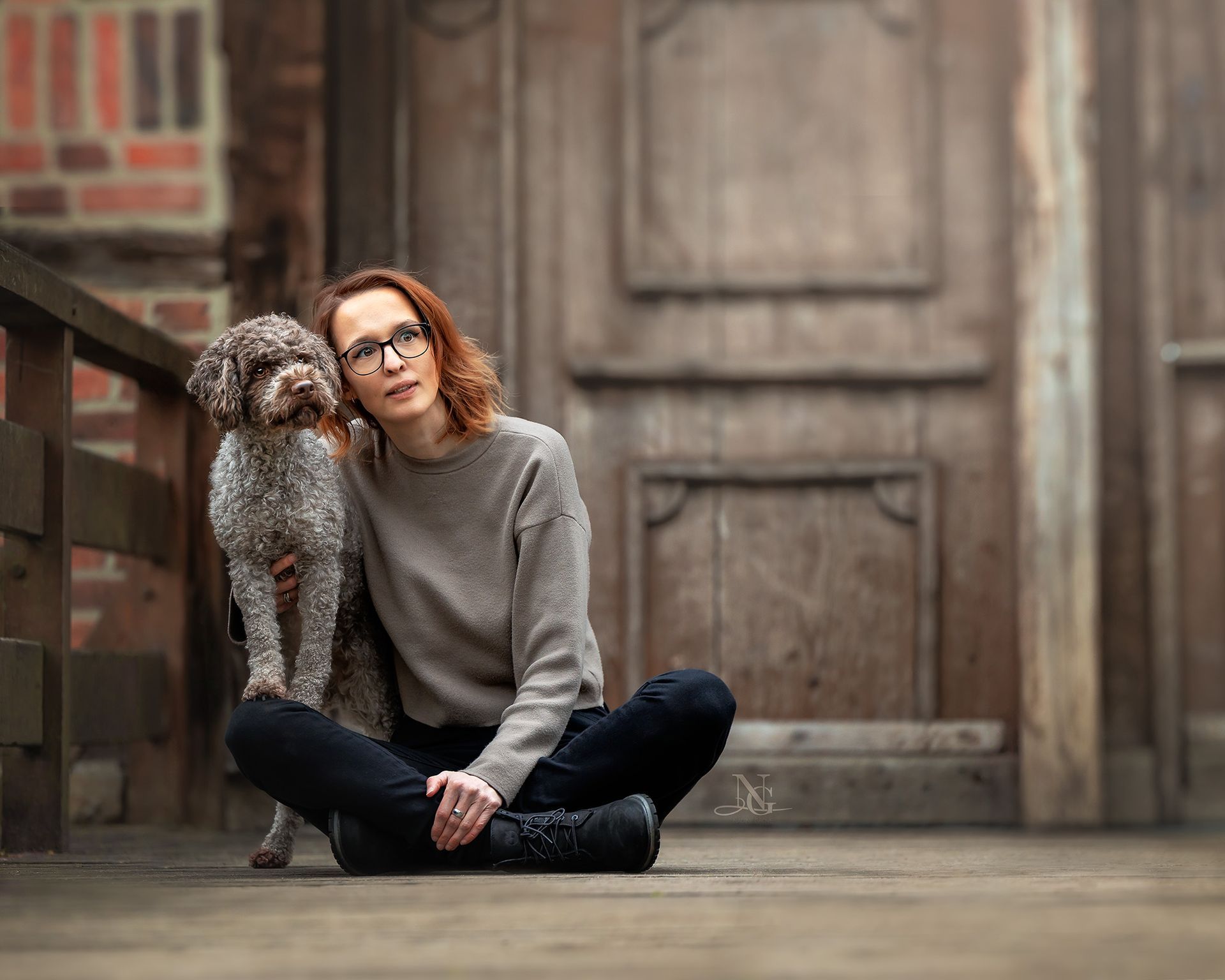 Frau sitzt im Schneidersitz auf dem Boden, Lagotto steht mit den Vorderpfoten auf dem Bein. Beide schauen in die Ferne.