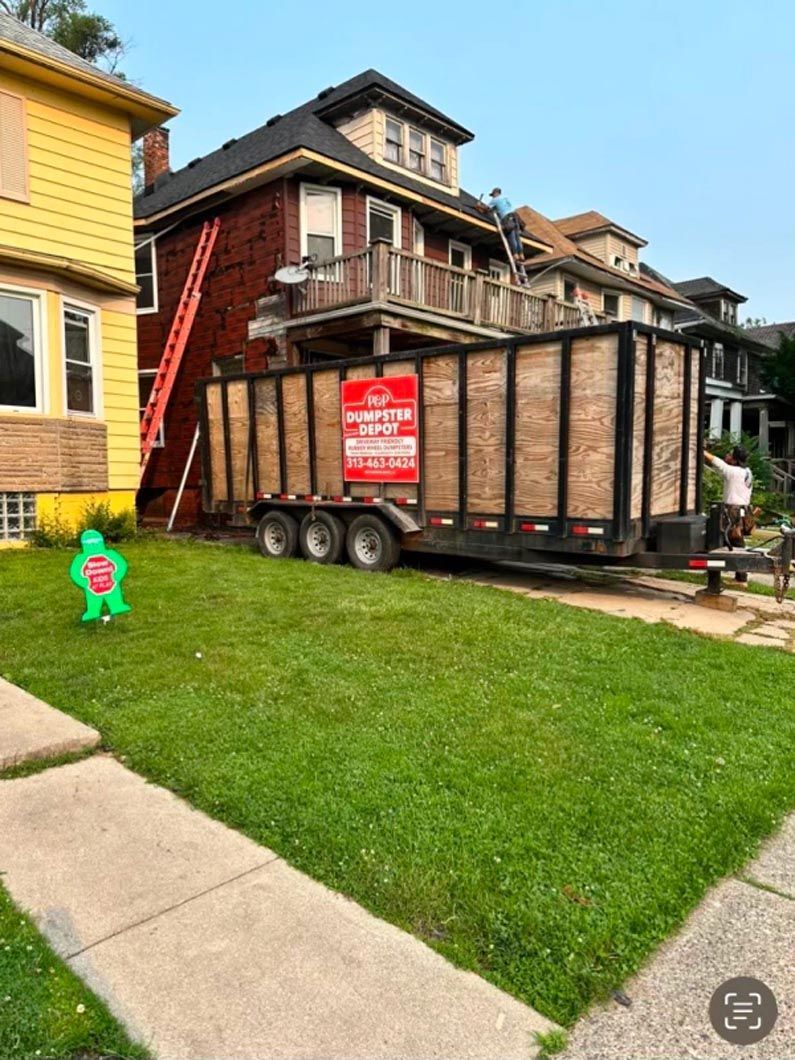 Large trailer parked on grass, next to a brick house. Orange ladder against the house. People on the balcony.