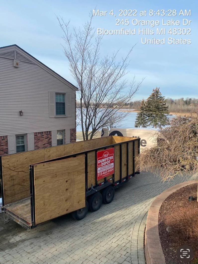 Trailer filled with tree branches parked in front of a beige building near a lake.