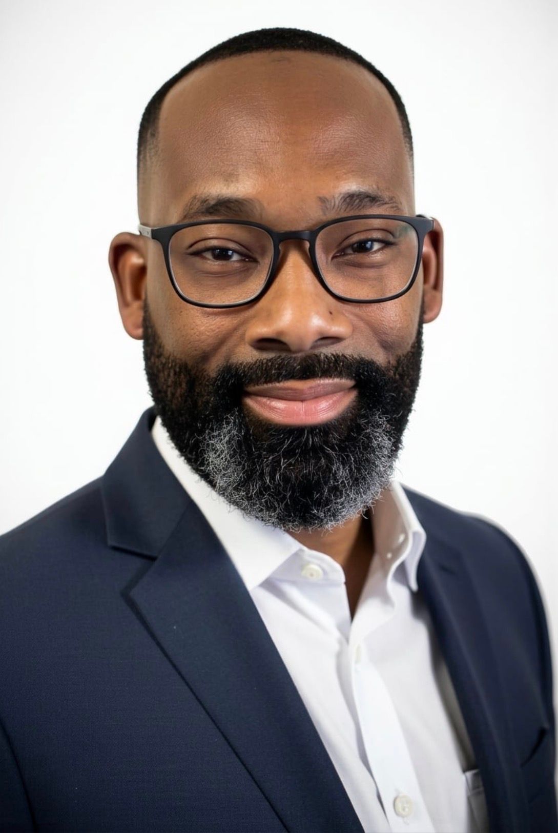Man with dark skin, neatly trimmed beard, in a suit, smiling, against a gray background.