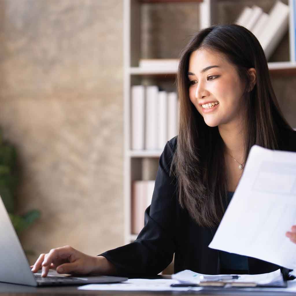 Woman smiles while working on a laptop, holding documents, in an office setting.