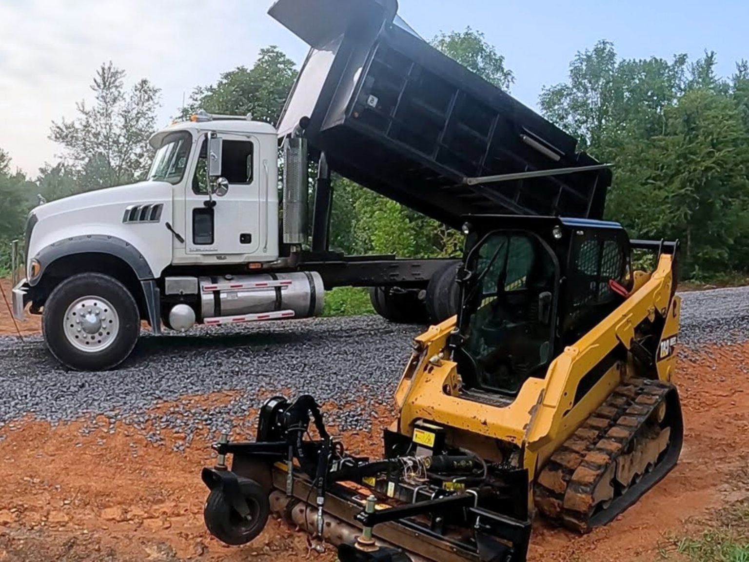 A dump truck unloads fresh gravel while a compact track loader levels the surface during a gravel driveway installation in Davenport.