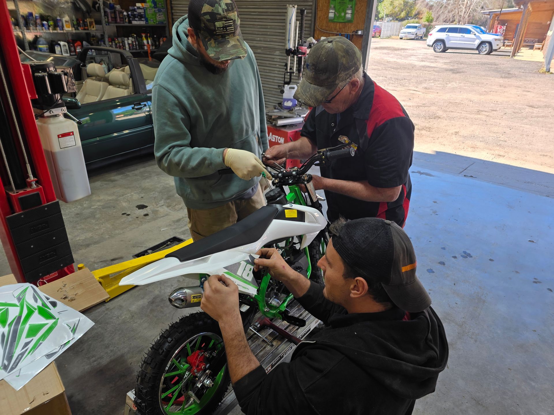 Three people work on a white dirt bike in a garage, with tools and parts spread on a table. | Masters Auto Group