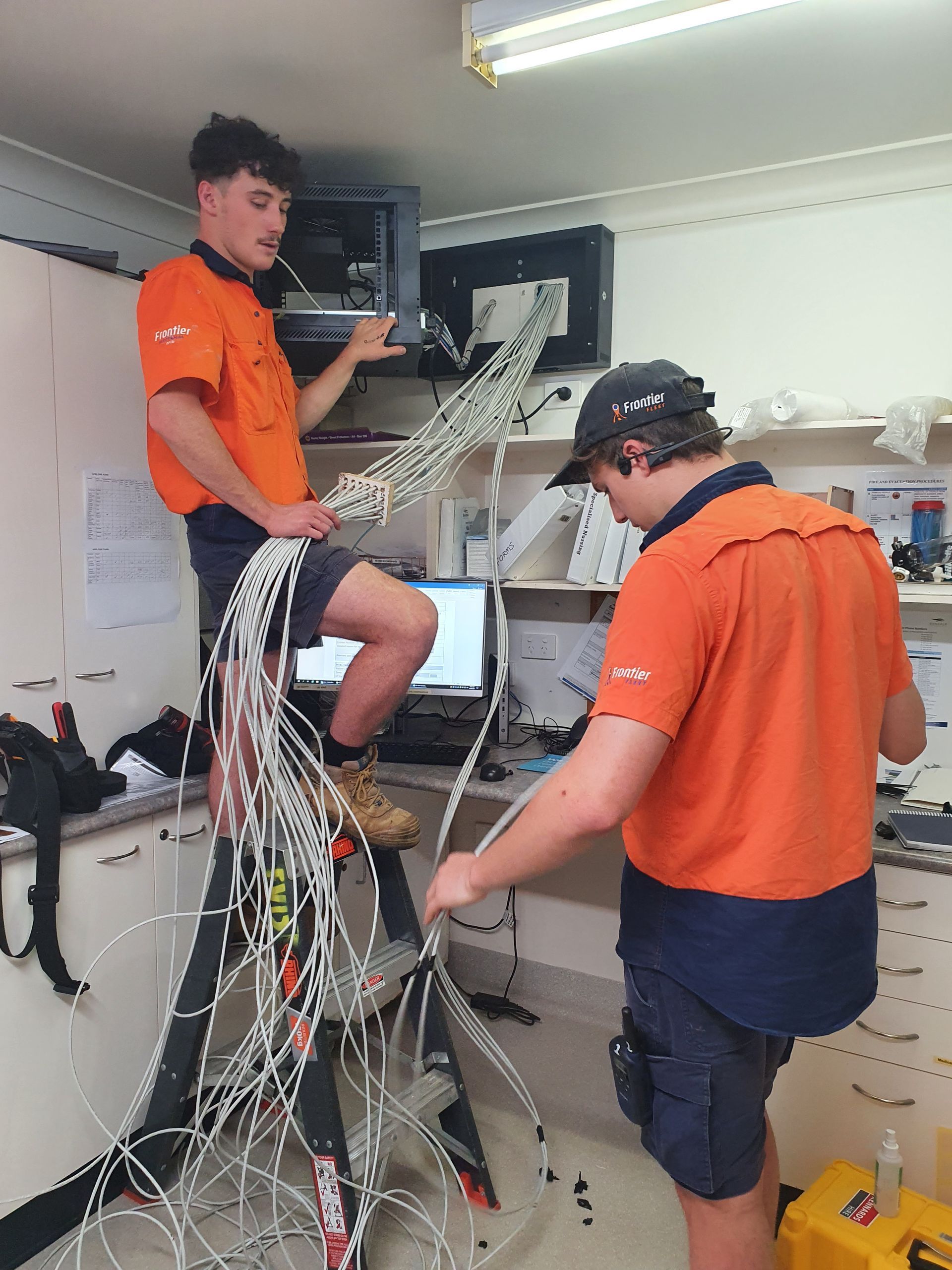 Two Men in Orange Shirts Work With Tangled Wires in an Office. One Stands on a Ladder — Frontier Security in Gatton, QLD