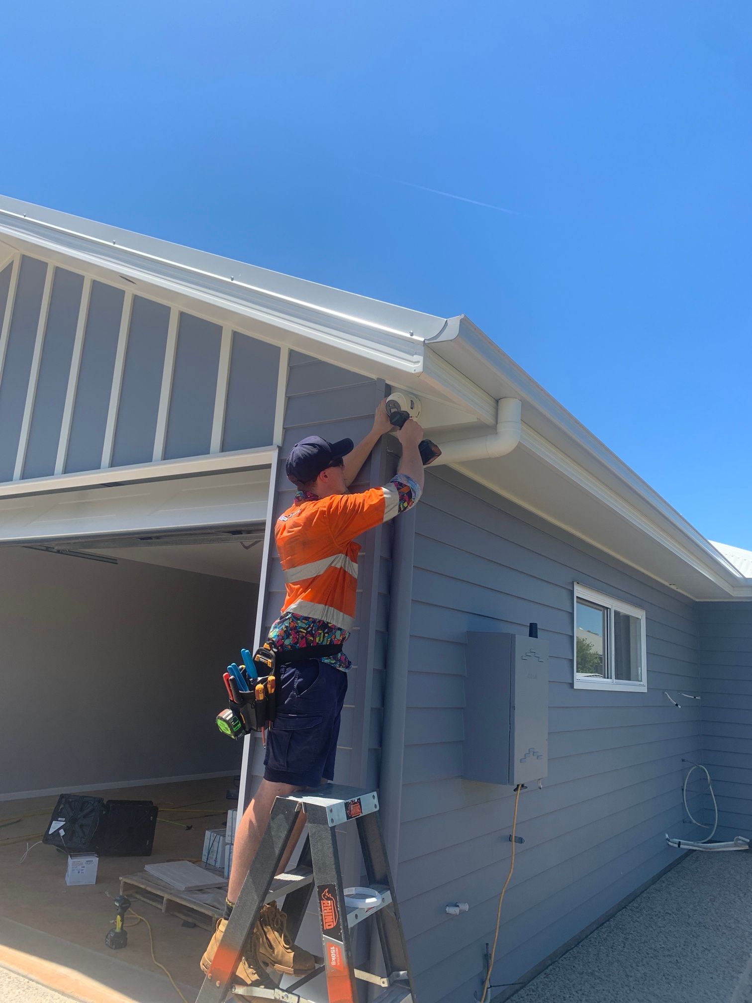 Man on Ladder Installing Gutter on a Gray Building Under a Blue Sky — Frontier Security in Chinchilla, QLD