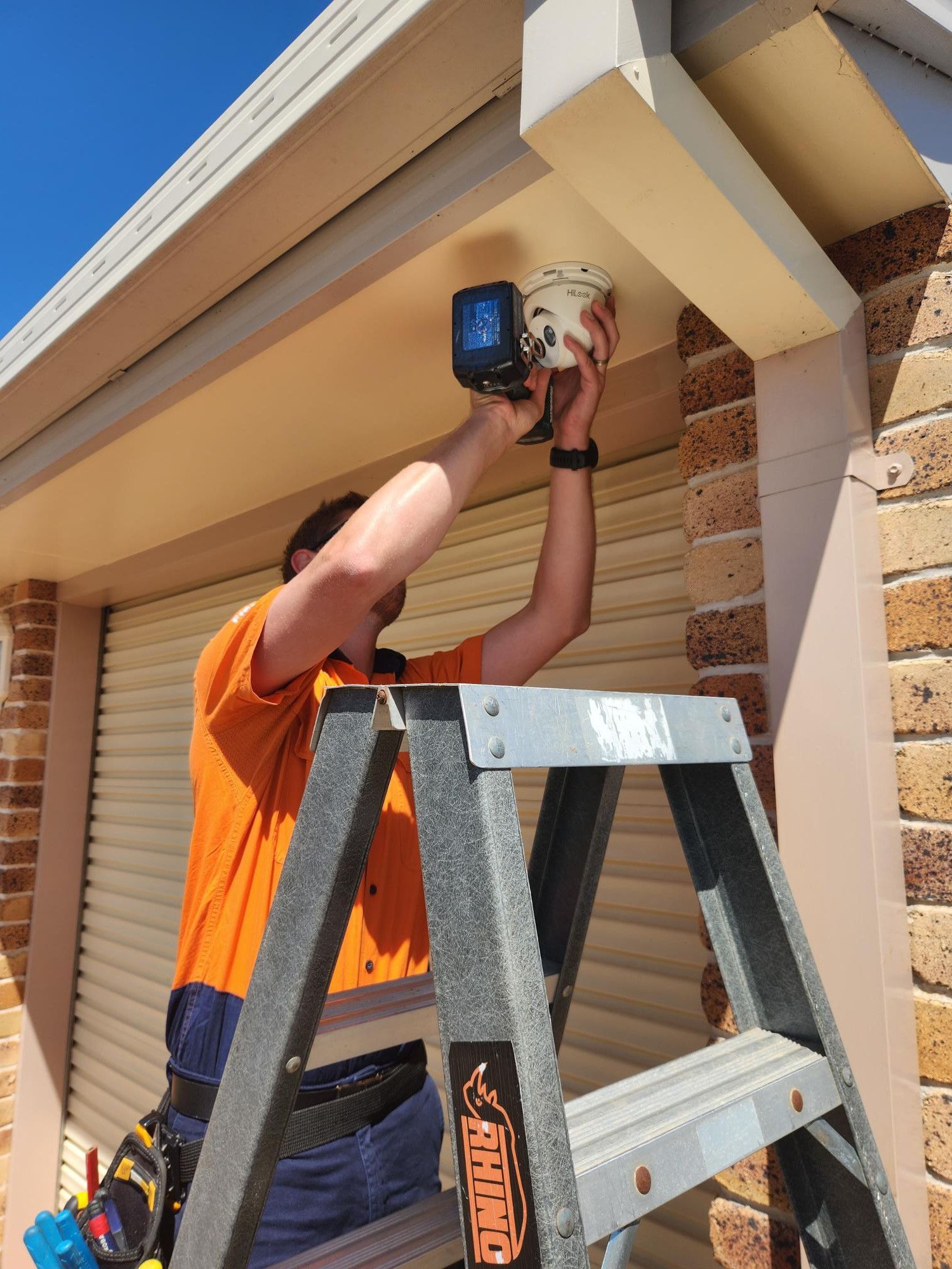 Man in Orange Shirt on Ladder Installing a Security Camera Under a Beige Awning — Frontier Security in Dalby, QLD