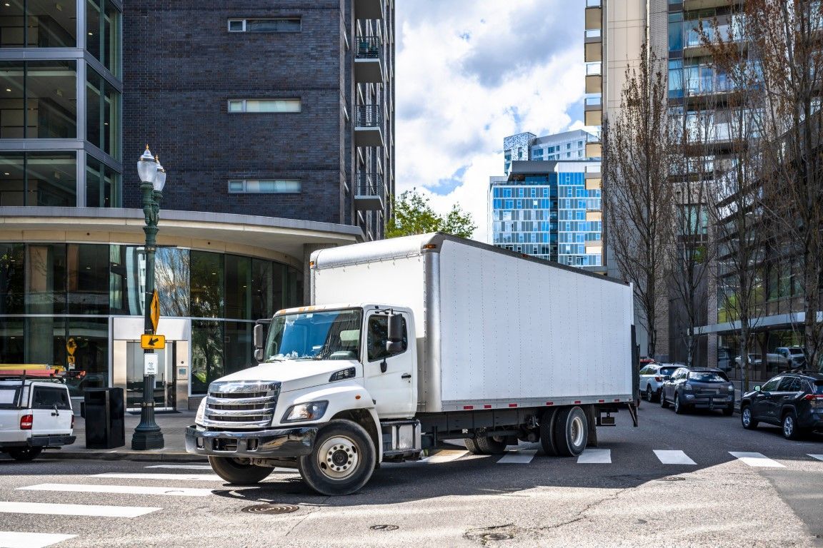 White box truck turning on a city street beside tall buildings and parked cars