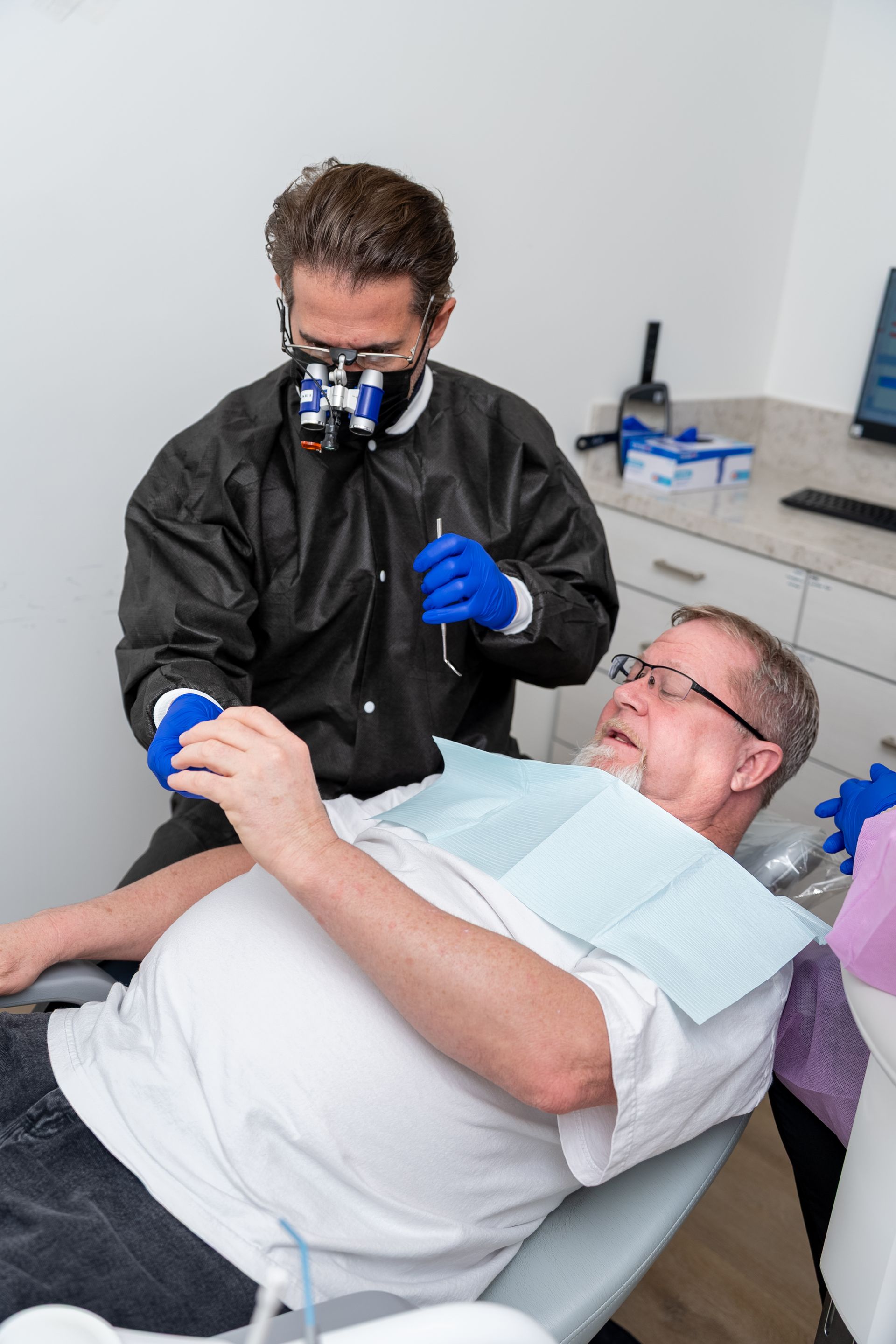 Dentist, in scrubs and loupes, examines a patient in a dental chair. Patient appears anxious.