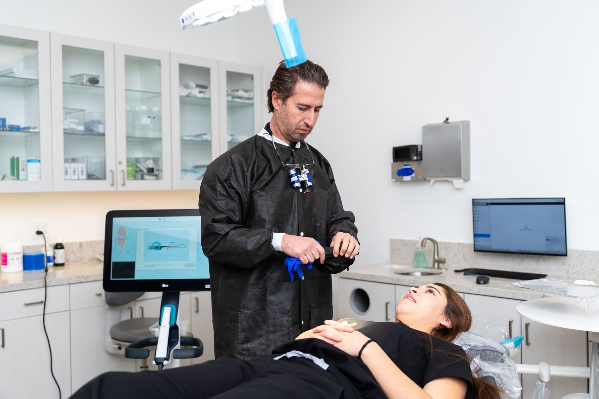 Dentist in black gown puts on gloves, attending to patient in dental chair, modern office.