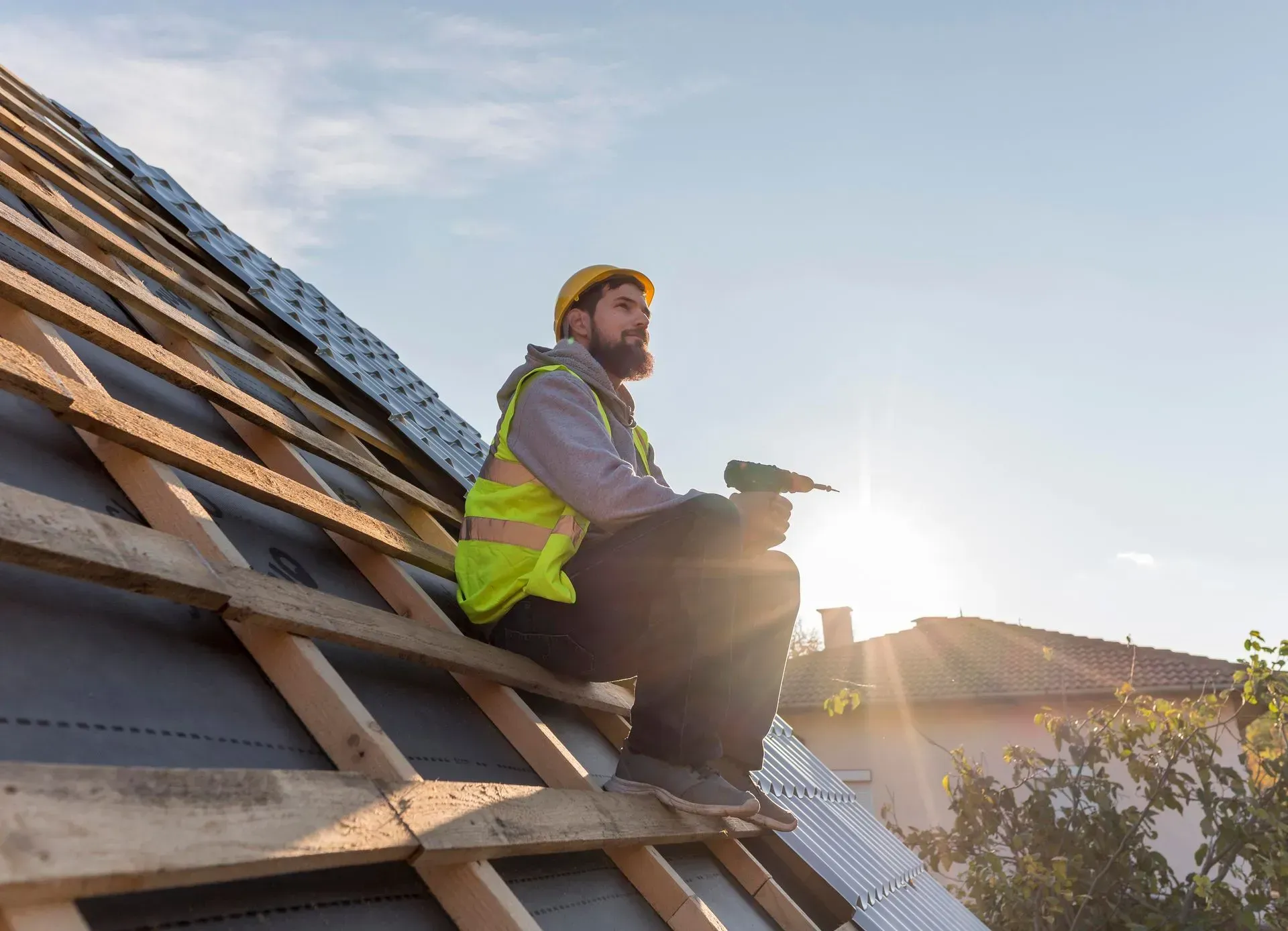 Roofer in safety vest and hard hat sits on roof, holding a tool, working on shingles in the sunlight.